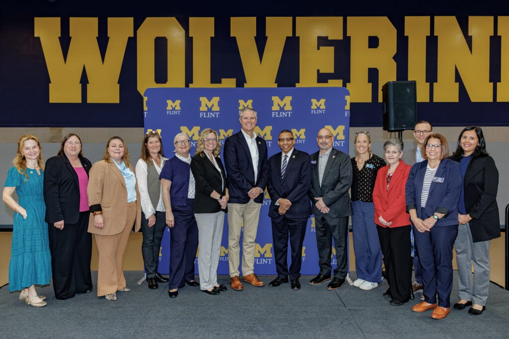A group of diverse individuals poses for a photo in front of a blue and yellow backdrop that reads "M FLINT" and "WOLVERINES."