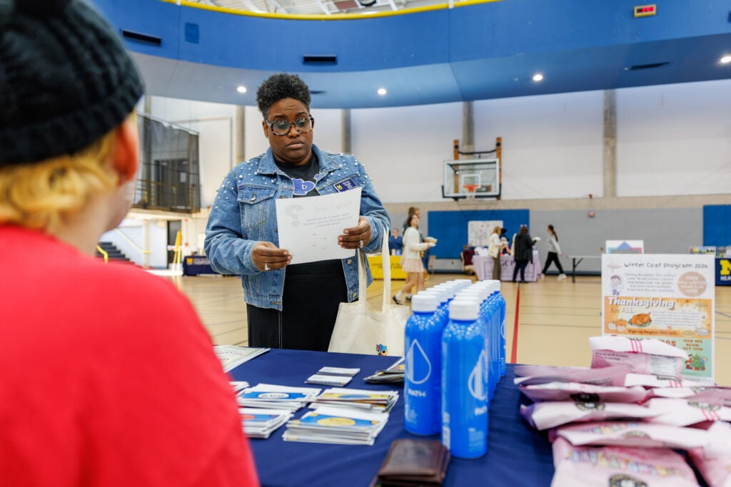 A person with glasses and a denim jacket is looking at a document while standing at a table with brochures and water bottles. In the background, people are engaging in activities in a gymnasium.