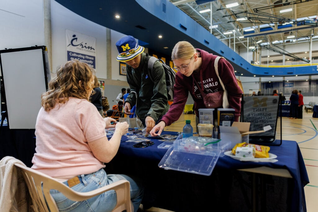 Two students are interacting at a table in a gymnasium, examining items laid out on a blue tablecloth. One student, wearing a green jacket and a hat, is pointing at the table, while the other, in a maroon sweatshirt, is looking closely at the items. A woman with curly hair is seated at the table, also engaged in the activity.