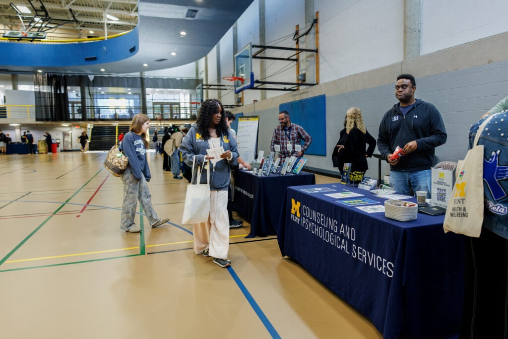 A gymnasium setting with people interacting at tables. A woman in a gray sweatshirt examines materials at a table labeled "M FLINT COUNSELING AND PSYCHOLOGICAL SERVICES." Other attendees browse nearby tables.