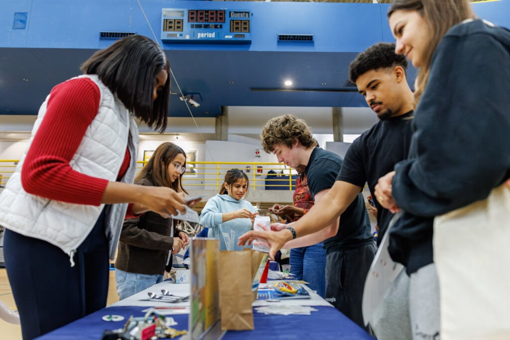 A group of five people are engaged in an activity at a table in a gymnasium, with various items spread out in front of them. A scoreboard is visible in the background.