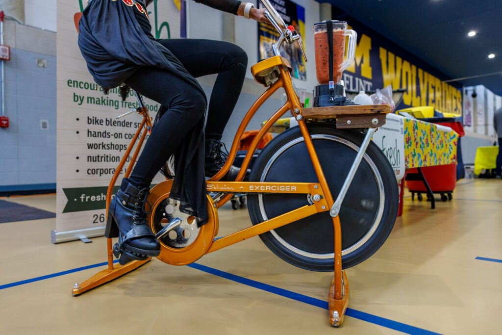 A person in black clothing is pedaling an orange stationary bike with a blender on top, blending a smoothie. In the background, there is a banner promoting fresh food initiatives.