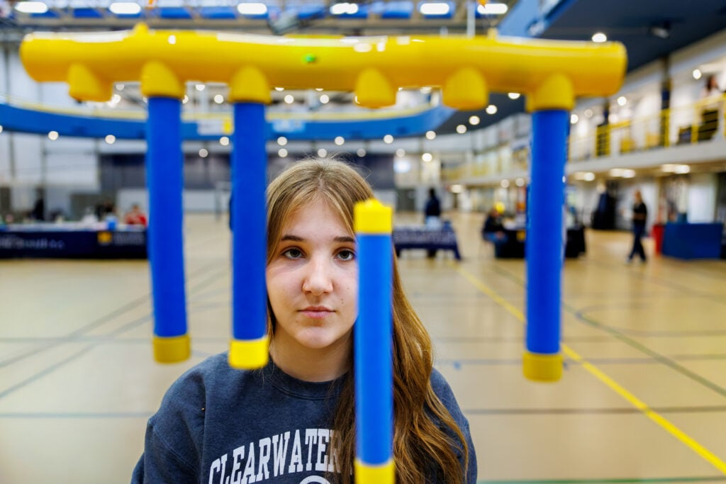 A young woman with long hair is looking directly at the camera, framed by colorful blue and yellow structures in a gymnasium setting.