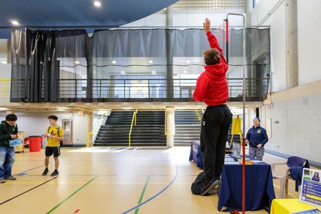 A person in a red hoodie jumps to reach a measuring device in a gym, while two others observe nearby.