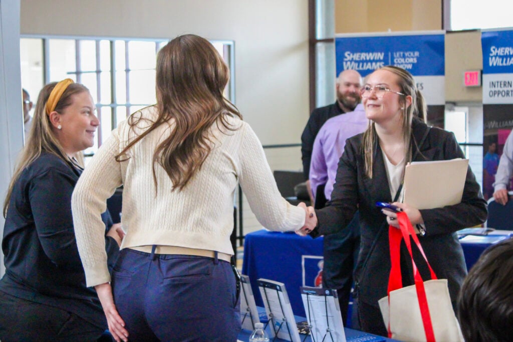 A job fair scene with two women shaking hands, one in a white sweater and the other in a black suit, while a third woman smiles nearby. Background features a Sherwin-Williams booth.
