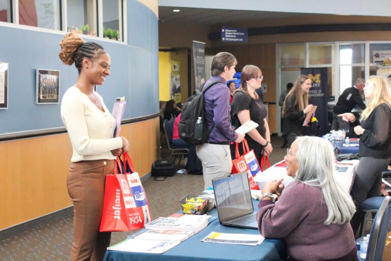 A young woman with braided hair smiles while holding a red bag and talking to a table representative at a career fair. Other attendees are visible in the background.