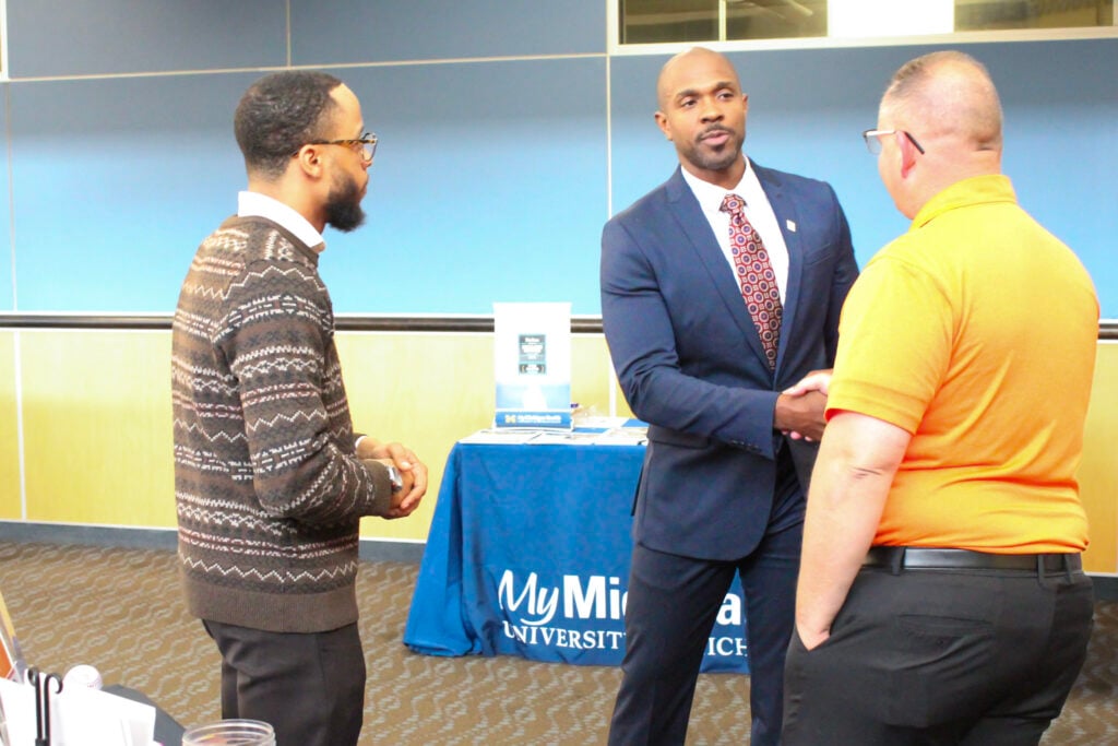 Three men are engaged in conversation at a table with a blue tablecloth featuring "My Minnesota University." One man in a suit is shaking hands with another man in an orange shirt, while a third man in a patterned sweater looks on.