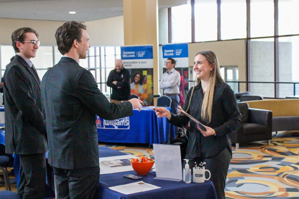 A woman in a black suit hands a document to a man in a suit at a table with promotional materials, while another man observes.