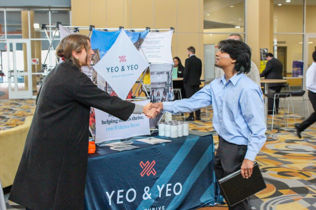 A woman in a black coat shakes hands with a man in a blue shirt at a Yeo & Yeo booth, with promotional materials displayed.