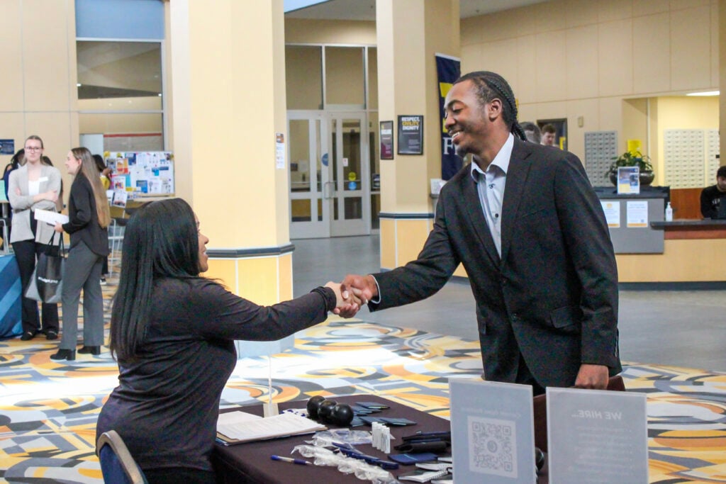 A man in a suit shakes hands with a woman at a table, while others engage in conversation in a busy indoor setting.
