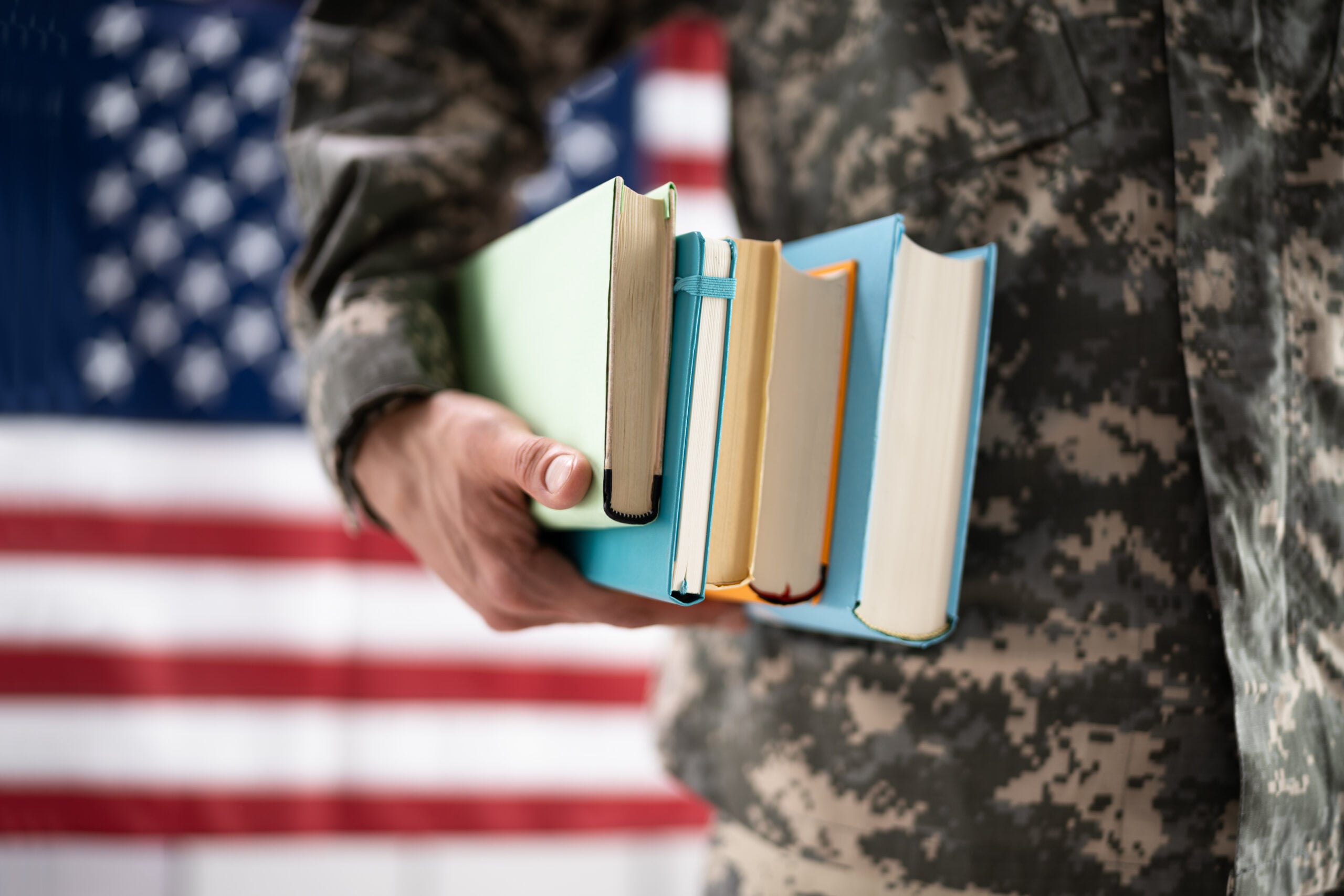 A person in military uniform holds a stack of colorful books in front of an American flag.