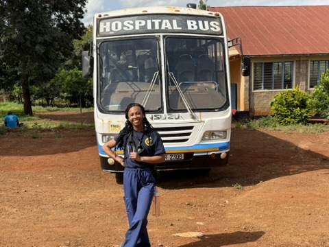 A smiling person in blue scrubs stands in front of a bus labeled "HOSPITAL BUS" in a rural setting.