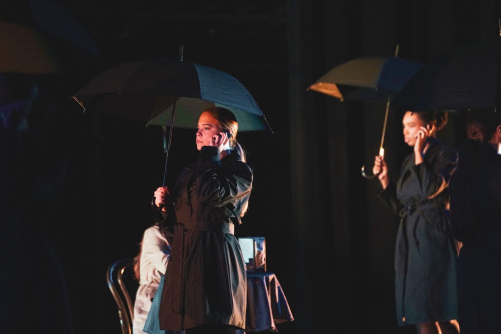 A theatrical performance featuring three women in dark coats holding black umbrellas. One woman is on the phone, while another stands nearby.
