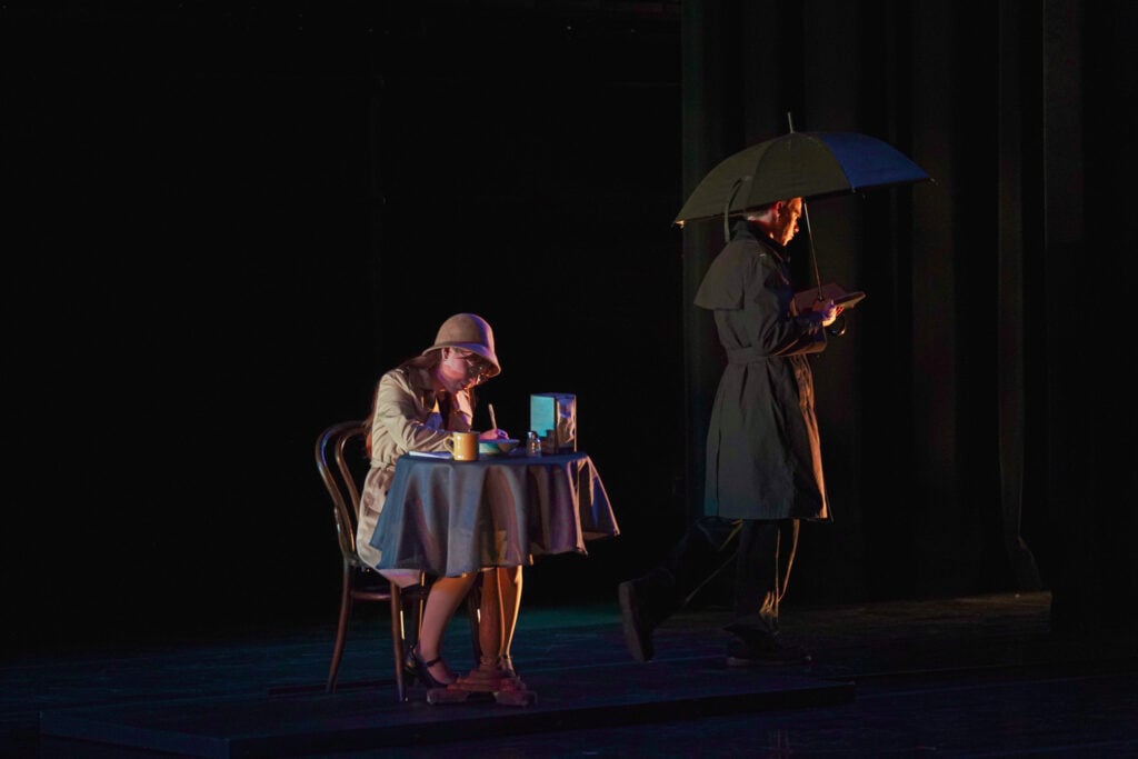 A woman in a hat sits at a table with a cup and a book, while a man in a trench coat walks by holding an umbrella.