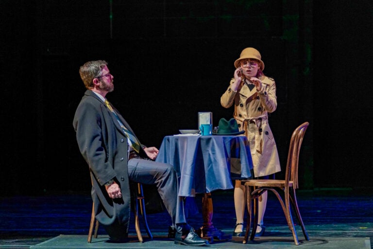 A man in a suit sits at a table, while a woman in a trench coat and hat stands nearby, appearing surprised or concerned.