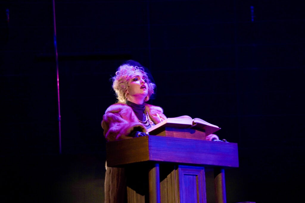 A performer in a dramatic pose stands at a wooden lectern with an open book, wearing a fur coat and dramatic makeup, under purple stage lighting.