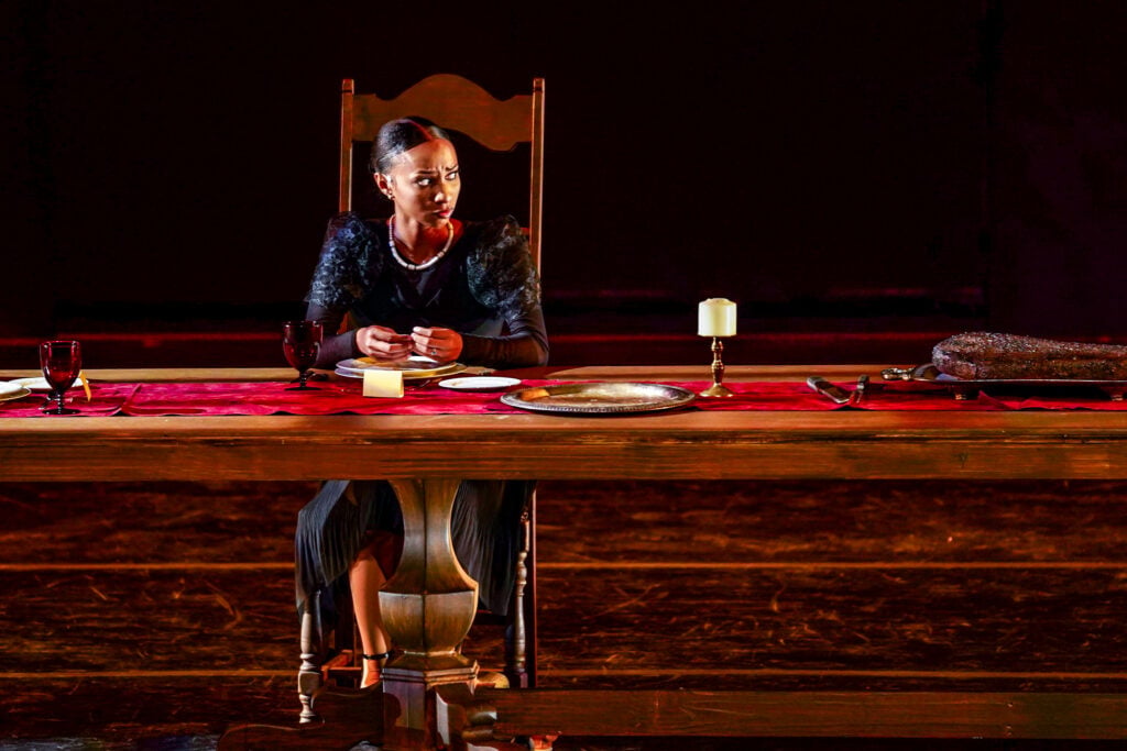 A woman sits at a long wooden table set with plates and glasses, looking thoughtfully at her hands. A candle and a large piece of food are on the table. The scene is dimly lit, creating a dramatic atmosphere.