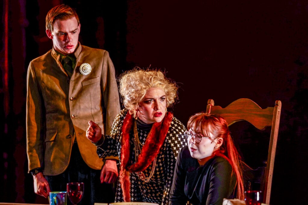 A dramatic scene from a play featuring three characters: a man in a suit looking concerned, a woman in a patterned outfit gesturing emphatically, and a young woman in glasses appearing distressed.