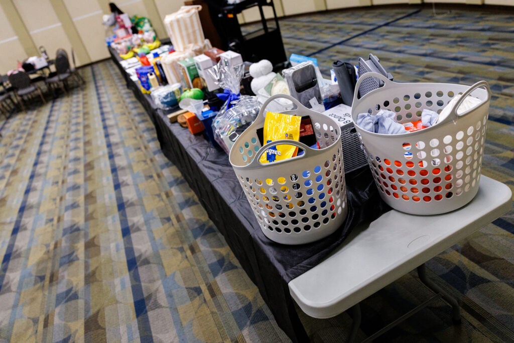 Two gray baskets filled with various items sit on a table, with a long display of products in the background. The setting appears to be an indoor event space with a patterned carpet.