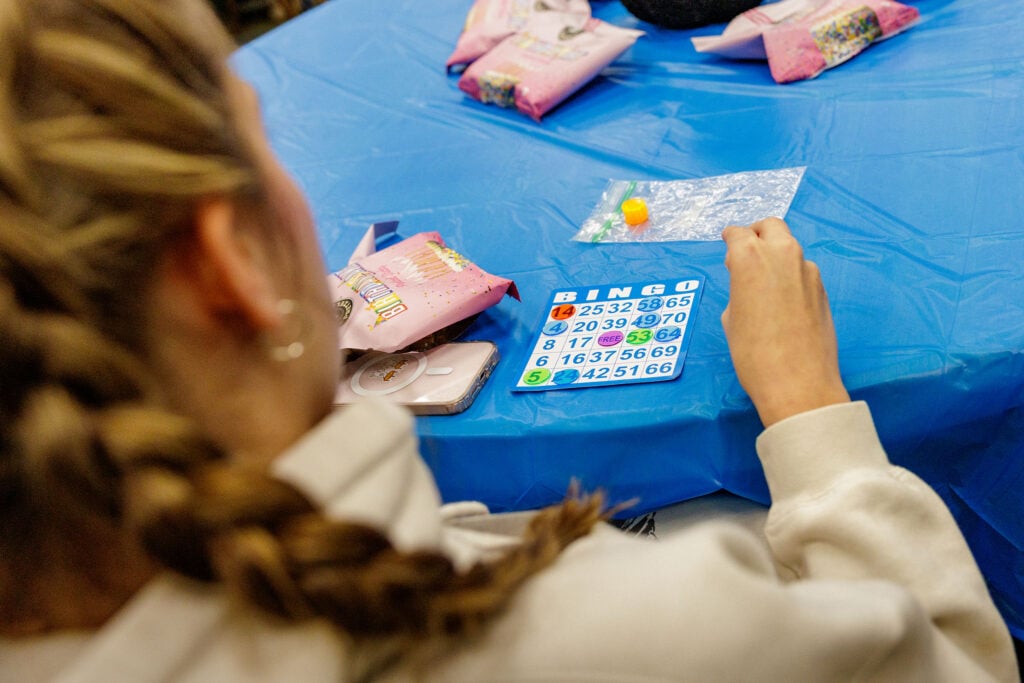 A person with braided hair is playing bingo at a table covered with a blue tablecloth, holding a bingo card with colorful numbers and a marker.
