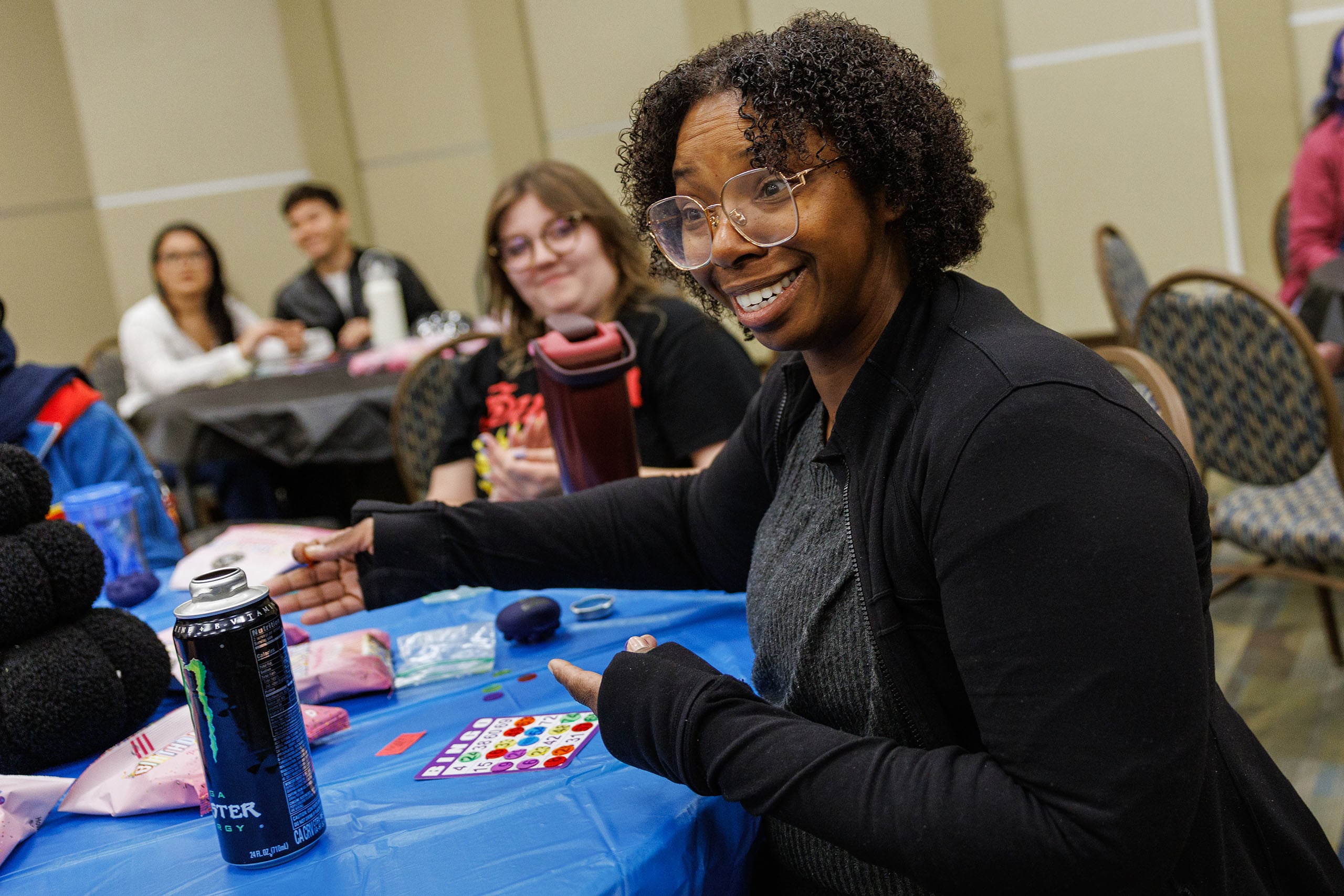A smiling person with curly hair and glasses sits at a table covered in a blue cloth, pointing at a bingo card. A can of Monster energy drink is in front of them, along with other game materials. In the background, other people are engaged in conversation.