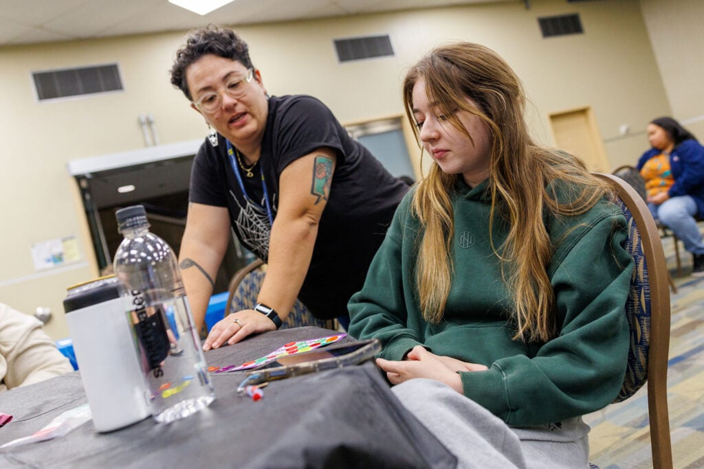 A person with short, curly hair and glasses is speaking to a young woman with long hair, who is sitting at a table. The table has a water bottle, a white container, and colorful stickers. Other people are visible in the background.