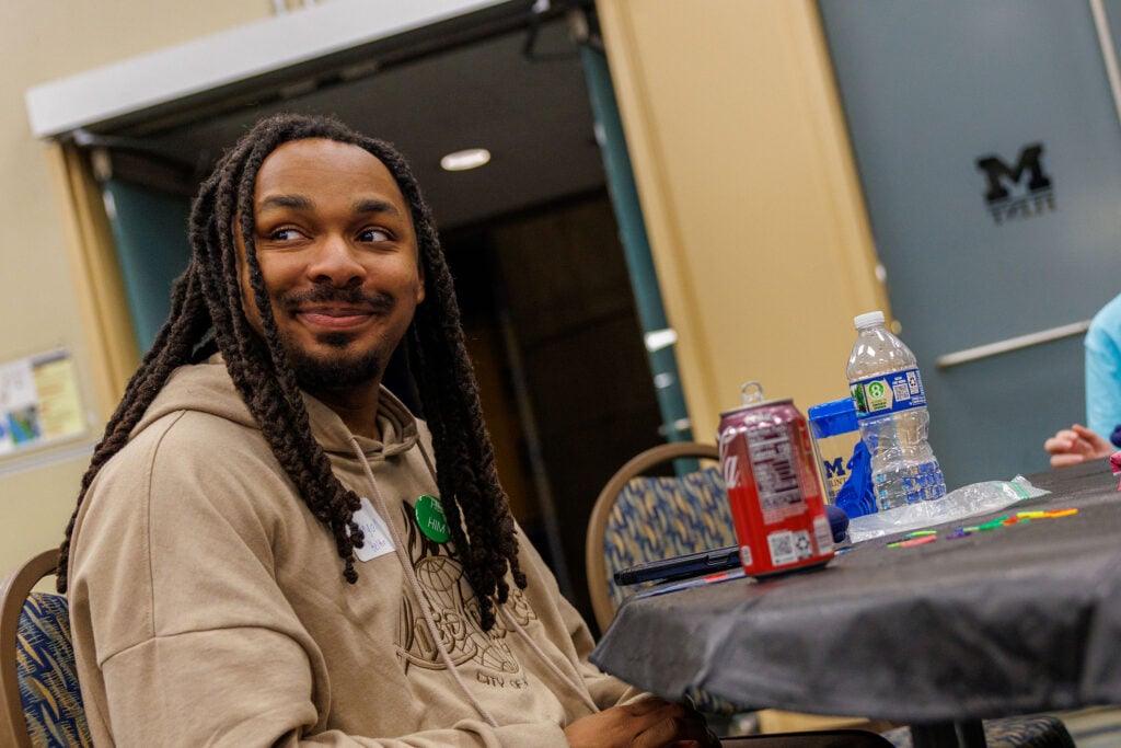 A person with long, curly hair and a light brown hoodie smiles while sitting at a table with drinks and colorful objects.