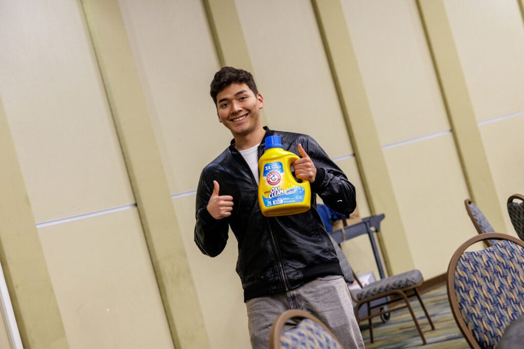 A young man in a black jacket smiles and gives a thumbs up while holding a bottle of laundry detergent in a room with chairs.