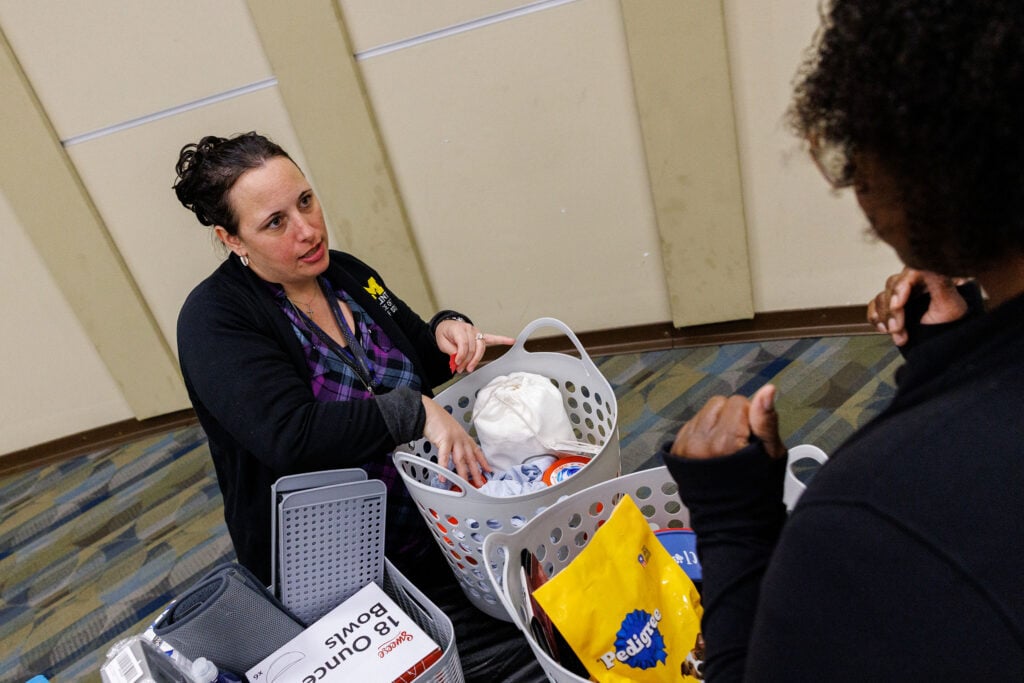 A woman with dark hair in a bun is speaking to another person while sorting items in a basket. The setting appears to be an indoor space with a patterned floor.
