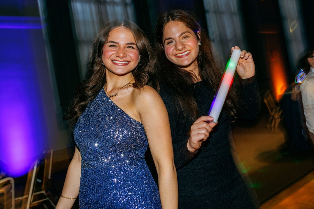 Two smiling women pose together at a party. One wears a sparkling blue dress, while the other holds a colorful light stick.