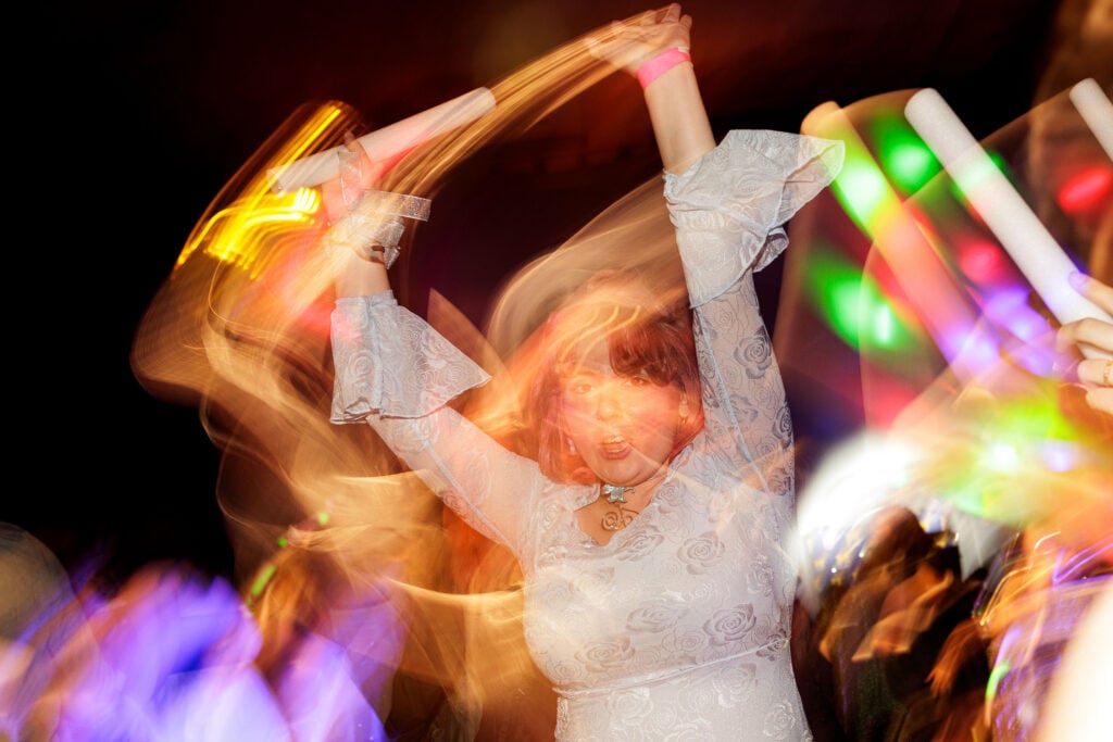 A woman in a white dress with bell sleeves raises her arms, holding light sticks, amidst colorful lights and motion blur, suggesting a lively party atmosphere.