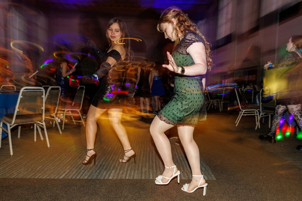 Two women dance in a lively setting, wearing stylish dresses and high heels, with colorful lights creating a dynamic atmosphere.