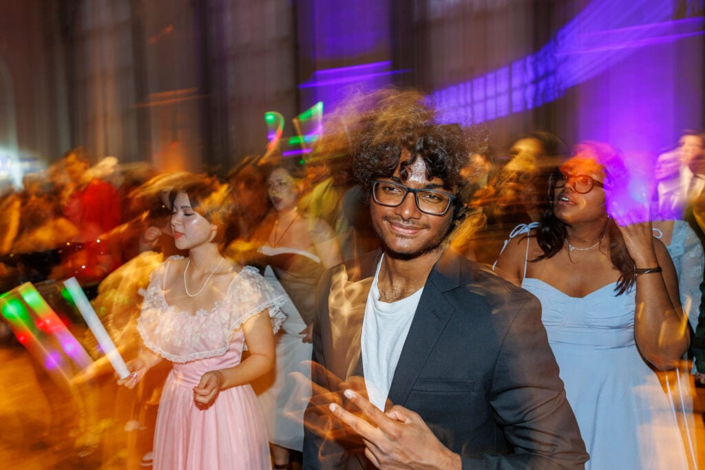 A young man in a black blazer smiles and makes a peace sign, surrounded by blurred figures dancing at a lively event.