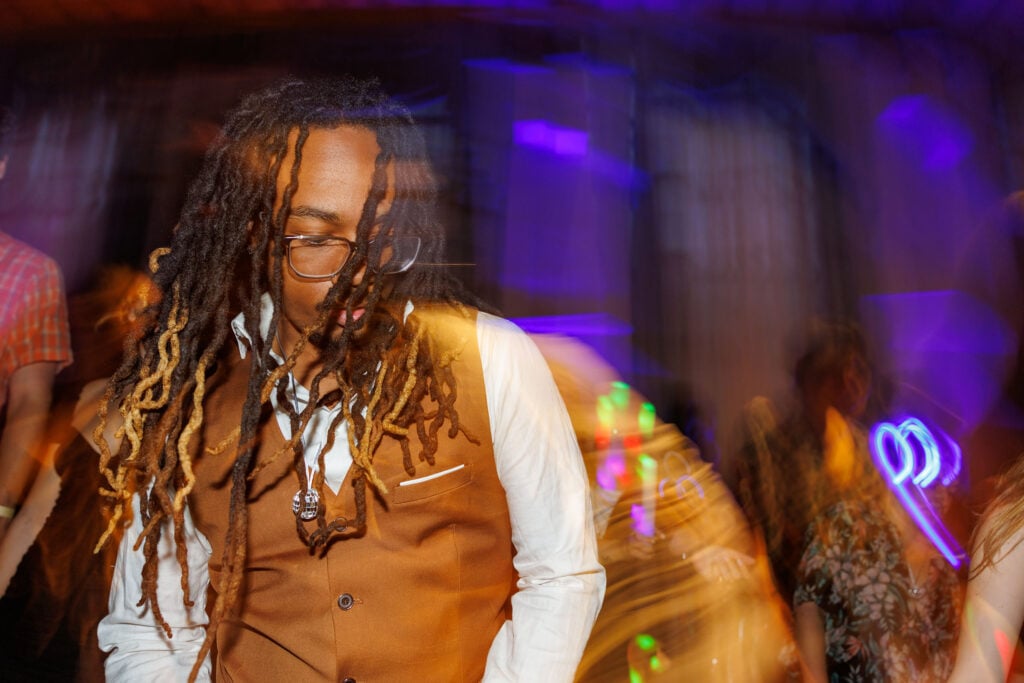 A person with long, curly hair wearing a brown vest and white shirt is dancing in a colorful, blurred party scene.