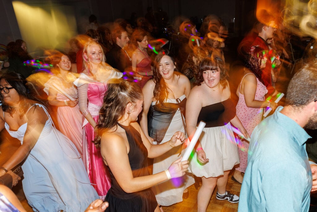 A lively dance floor scene with a group of young people in formal attire, enjoying a celebration with colorful lights and glow sticks.