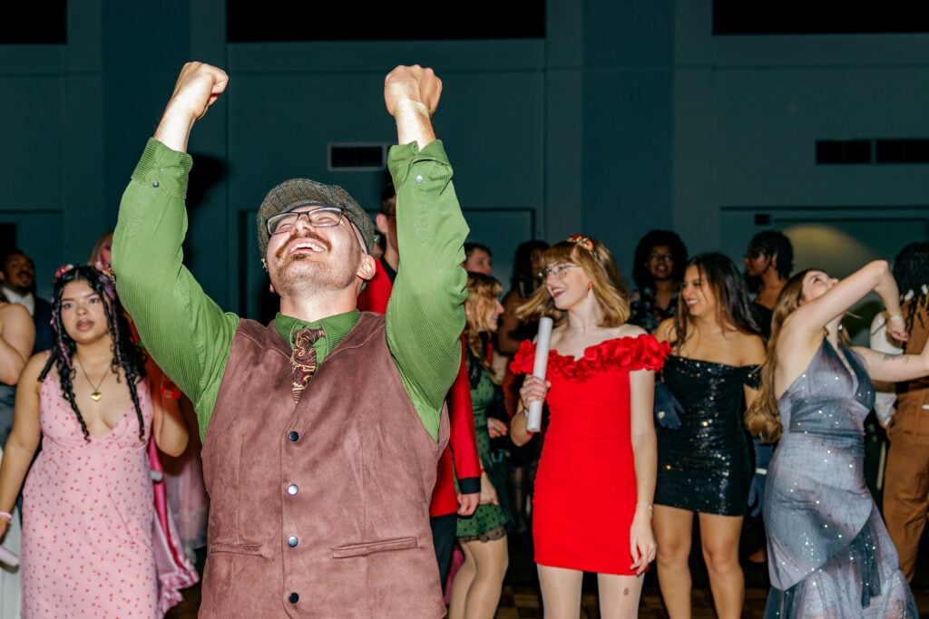 A group of people dancing at a party, with a man in a green shirt and brown vest raising his arms in celebration.