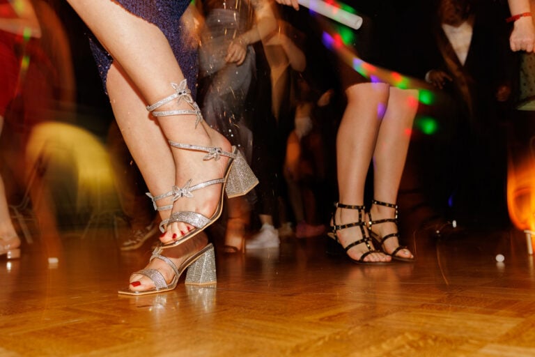 A close-up of two pairs of women's shoes on a dance floor, one pair is silver strappy heels and the other is black strappy sandals.