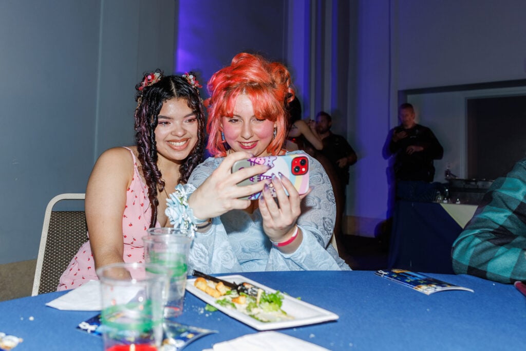 Two young women are sitting at a table, smiling and taking a selfie with a phone. They are dressed in colorful outfits, with one having pink hair and the other with dark, curly hair. A plate of food and drinks are on the table in front of them.