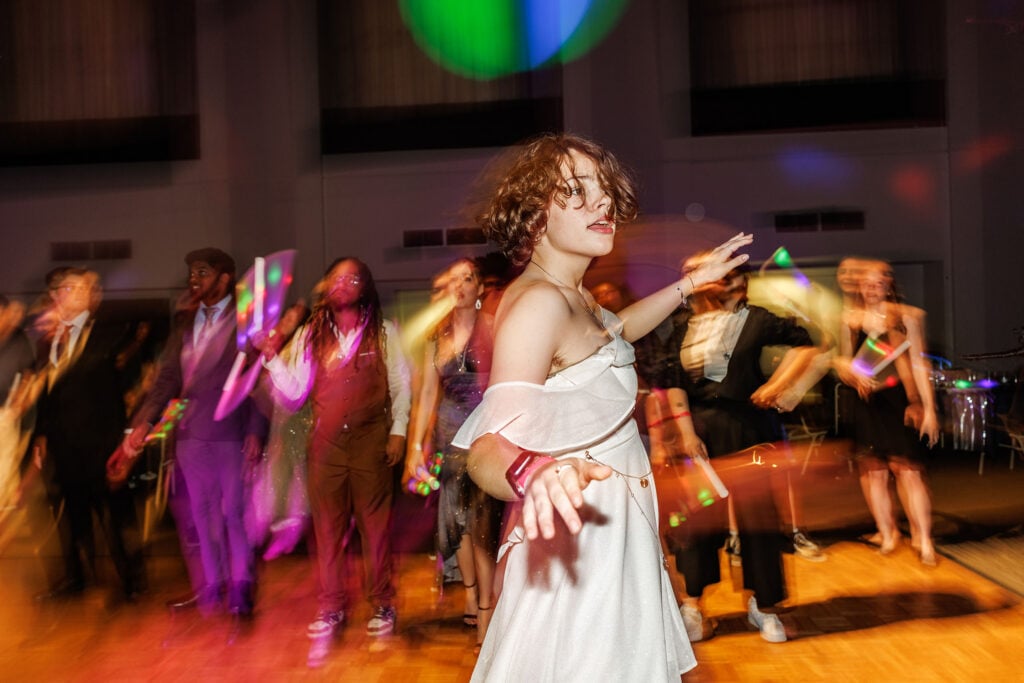A group of people dancing at a lively event, with a focus on a young woman in a white dress, moving joyfully amidst colorful lights.