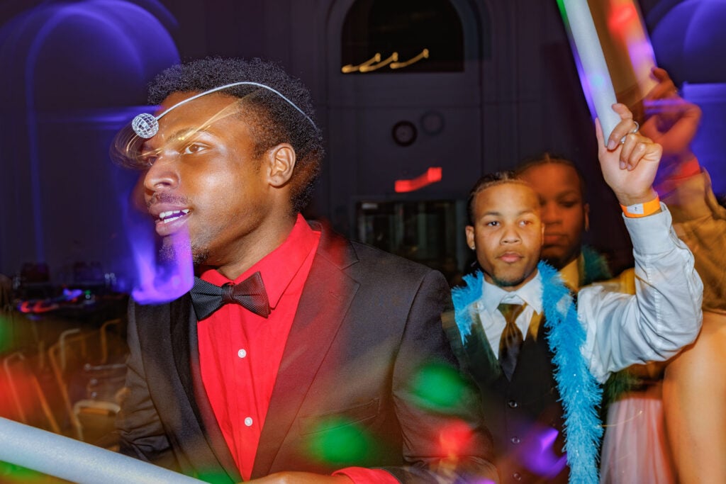 A man in a black suit and red shirt is smiling, wearing a decorative headpiece, while another man in a blue feather boa stands behind him, raising his hand. The background features colorful lights and a festive atmosphere.