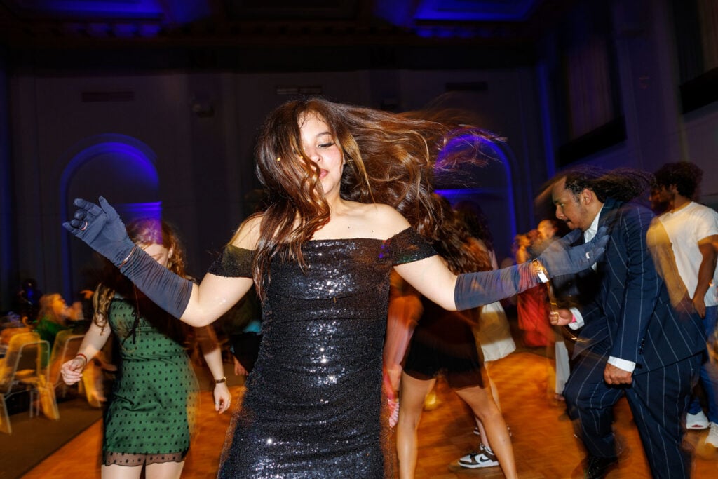 A woman in a sparkling black dress dances energetically, her hair flowing, with other people dancing in a lively setting.