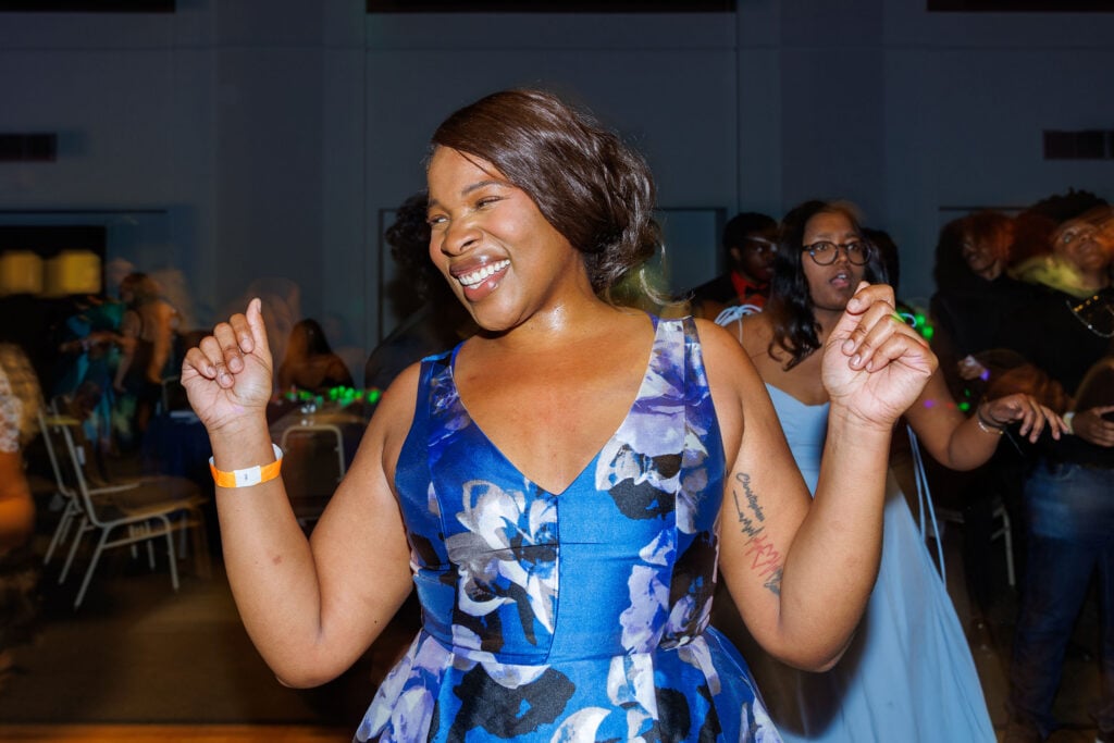 A woman in a blue floral dress smiles and dances, raising her hands in a lively atmosphere with other people in the background.