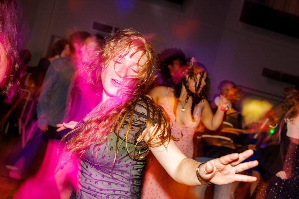 A woman with long, wavy hair dances joyfully in a vibrant, colorful setting, surrounded by other partygoers.