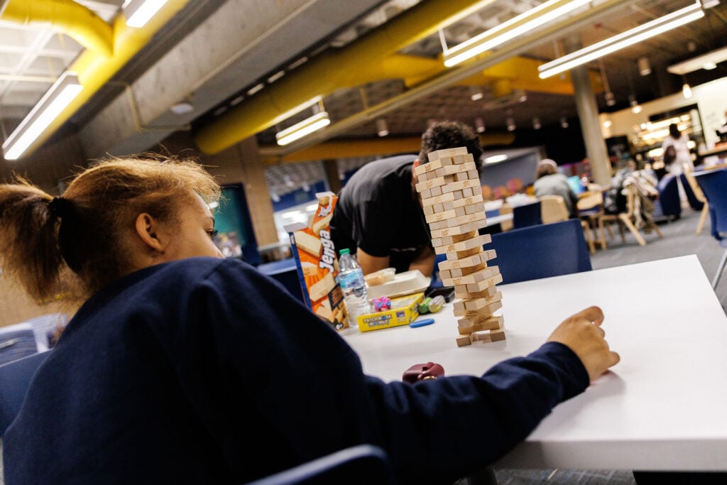 A child with curly hair is focused on a Jenga tower made of wooden blocks on a table, while another person is in the background.