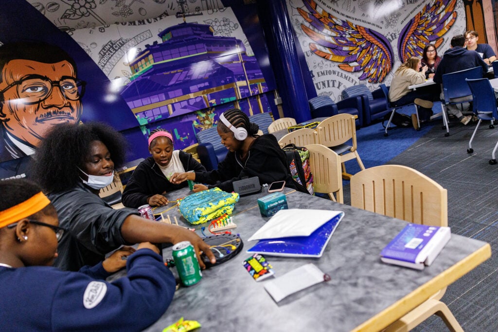 A group of four young people gather around a table with a colorful cake, snacks, and drinks, while a mural of a man with glasses is visible in the background.