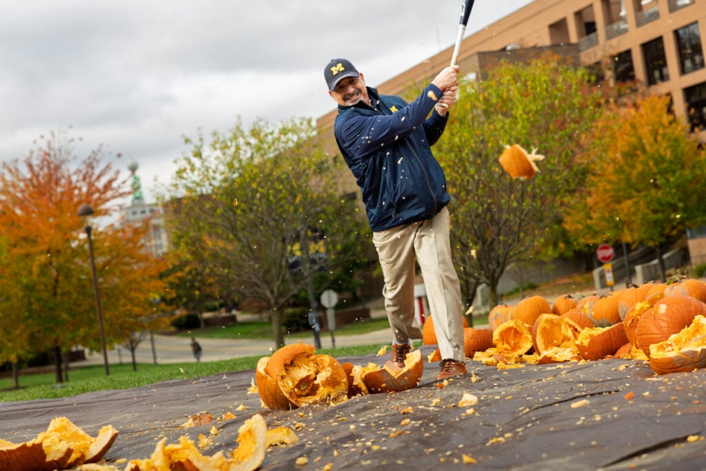 A man in a blue jacket and cap swings a bat at a pile of smashed pumpkins, with pumpkin guts flying in the air. Autumn trees are in the background.