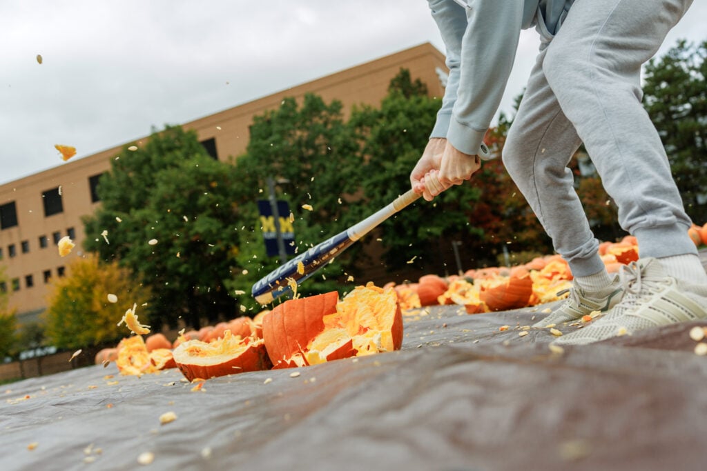 A person in gray sweatpants and a hoodie swings a bat at a pumpkin, causing pumpkin guts to fly in the air. Many smashed pumpkins are scattered on the ground.