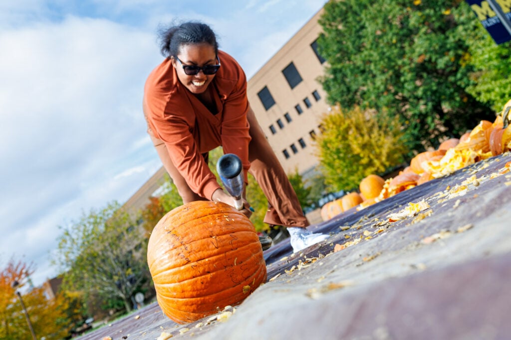 A person in sunglasses is lifting a large pumpkin while standing on a path surrounded by other pumpkins and autumn foliage.
