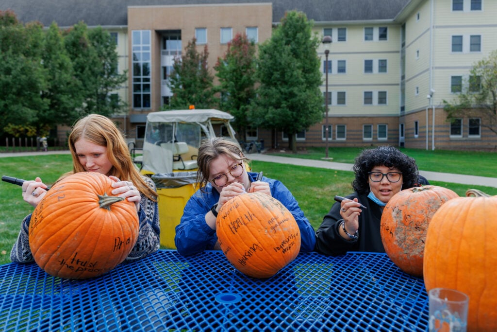 Three people are sitting at a blue table, decorating pumpkins with markers. They are focused on their work, with several pumpkins in front of them. In the background, there are trees and a building.