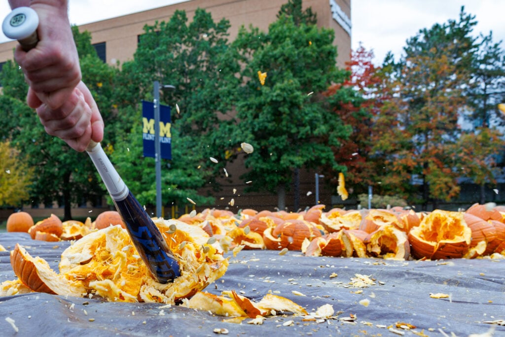 A person swings a bat to smash a pumpkin, with pumpkin pieces flying in the air. Many carved pumpkins are scattered on the ground.
